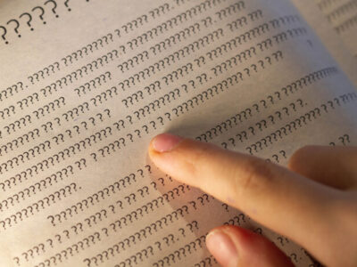 A person leads a finger on the lines in the book, but instead of letters only question marks on the page in the textbook